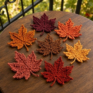 Può includere: Una collezione di sette foglie d'acero all'uncinetto in vari colori autunnali, tra cui bordeaux intenso, arancione brillante, giallo dorato, marrone scuro e rosso ruggine, ciascuna con punti dettagliati e una forma a punta distinta.