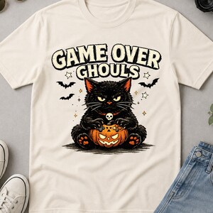 Op de afbeelding: Een crèmekleurig t-shirt met een afbeelding van een zwarte kat met een schedelketting die een pompoen vasthoudt. De tekst GAME OVER GHOULS staat erboven, vergezeld van kleine illustraties van vleermuizen en sterren op de borst.