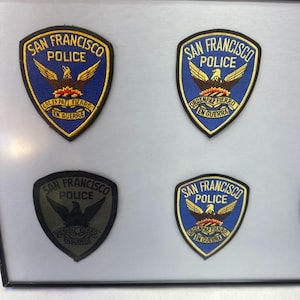 May include: Four shield-shaped San Francisco Police patches displayed in a frame. Three patches feature a blue background with a gold eagle and the text San Francisco Police and Orden Paz Fierro En Guerra. One patch is monochromatic dark gray.