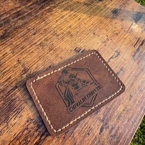 May include: A rectangular brown leather patch with rounded corners and light-colored perimeter stitching. The center features a black laser-engraved logo of a knight holding a hammer inside a hexagon, with the text CHIVALRY CRAFT CO. below.