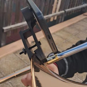 Op de afbeelding: Een zwarte smartphonehouder bevestigd aan de beker van een metalen trombone. De houder is voorzien van een schroefmechanisme en een plat oppervlak om het apparaat stevig op zijn plek te houden tijdens het bespelen van het instrument.