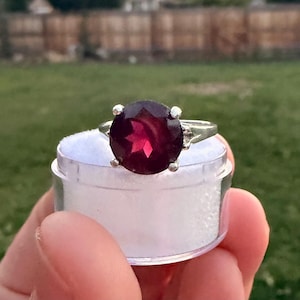 May include: A silver ring featuring a large, round, faceted deep red gemstone set in a four-prong mounting. The ring is positioned on top of a small, clear, cylindrical container filled with white material, showcasing the stone's dark red color.