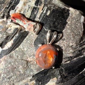 May include: A round, polished fire opal pendant set in a silver prong setting. The gemstone displays deep reddish-orange and brown hues with internal flashes of bright orange light. The pendant features a silver bail at the top.