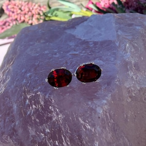 May include: A pair of oval-shaped stud earrings featuring deep red, faceted gemstones set in silver-toned metal prongs. The earrings are positioned on a large, translucent, light purple crystalline surface with a rough, jagged texture.