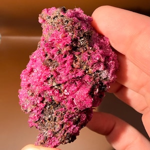 May include: A raw mineral specimen featuring a vibrant, saturated magenta-pink crystalline structure contrasted with dark, earthy brown and black matrix inclusions. The surface is highly textured, porous, and jagged, showcasing small, sparkling crystal facets.
