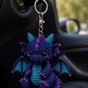 Könnte beinhalten: Ein gehäkelter Drachen-Schlüsselanhänger mit dunkelblauem Körper und hellen türkisfarbenen Akzenten an Bauch, Flügeln und Schwanz. Der Drache hat violette Stacheln am Rücken und Kopf, große schwarze Augen und einen silbernen Metallring.