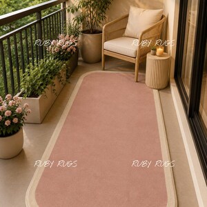 May include: A rectangular area rug with rounded corners, featuring a solid dusty pink center and a wide, light beige border. The text RUBY RUGS is visible in four locations on the rug and surrounding area. The rug has a flat, uniform texture.