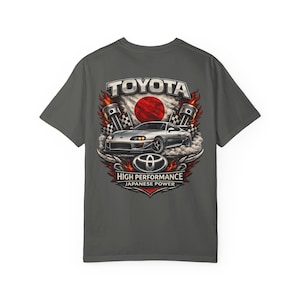 May include: A charcoal gray t-shirt featuring a graphic print of a silver sports car, the Toyota logo, and the text TOYOTA HIGH PERFORMANCE JAPANESE POWER. The design includes illustrations of engine pistons, checkered flags, flames, and a red sun.