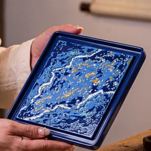 May include: A square ceramic tray with a deep blue border and a central decorative panel featuring an abstract pattern of swirling blue, white, and gold tones. The tray has a raised edge and a glossy finish, showcasing a complex, textured design.