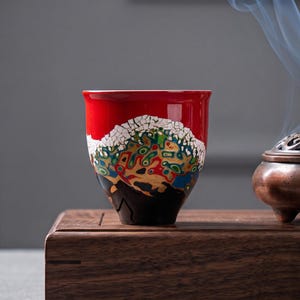 May include: A ceramic tea cup featuring a vibrant red upper section and a complex, multi-colored marbled pattern in the center, transitioning to a solid black base. The cup has a white crackle-glaze texture separating the red and marbled areas.