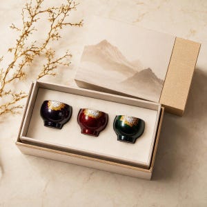May include: A set of three small, rounded cups in dark purple, deep red, and dark green, each featuring a gold-toned, textured rim. The cups are arranged inside a rectangular beige box with a lid depicting a mountain landscape illustration.