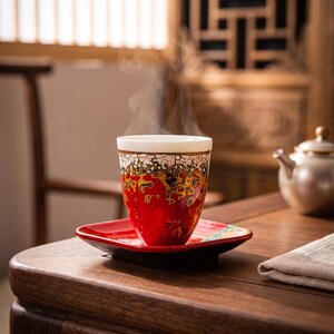 May include: A ceramic tea cup with a vibrant red, yellow, and black crackle glaze pattern sits on a matching square red saucer. Steam rises from the cup, which features a white interior rim. The set is displayed on a wooden surface.