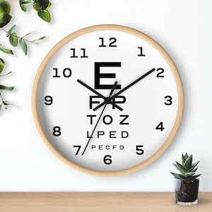 May include: A round wall clock with a light wood frame and a white face. The clock features black numbers from 1 to 12 around the perimeter and a central Snellen eye chart design with black letters E, FP, TOZ, LPED, and PECFD. It has black hands.