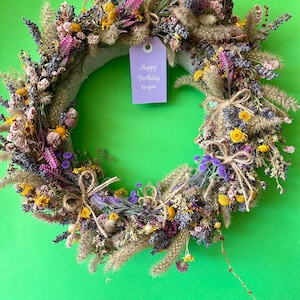 Könnte beinhalten: Ein runder Kranz aus getrockneten Blumen und Gräsern in Lila-, Gelb- und Grüntönen. Der Kranz ist mit kleinen Schleifen aus Jutegarn verziert und trägt ein hellviolettes, rechteckiges Schild mit der Aufschrift Happy Birthday to you.