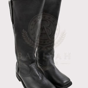 May include: A pair of tall black leather boots featuring a square toe design, a flat heel, and visible white stitching along the sole. The boots have a smooth finish and reach mid-calf height, with a structured shaft and a clean, simple silhouette.