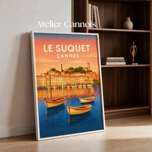 May include: A framed art print titled Atelier Cannois featuring a sunset illustration of Le Suquet in Cannes. The scene depicts a coastal town with buildings reflecting in blue water, accompanied by three small wooden boats floating in the foreground.