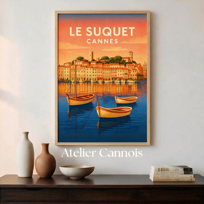 May include: A framed wall art print titled LE SUQUET CANNES featuring a coastal town with buildings in warm tones, a deep blue harbor, and three small yellow boats floating on the water under a sunset sky with orange and pink hues.