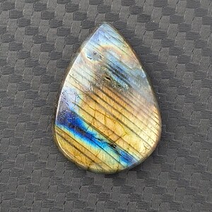 May include: A teardrop-shaped labradorite gemstone featuring a polished surface with iridescent flashes of blue, gold, and yellow. The stone displays natural internal striations and color variations across its smooth, rounded face.