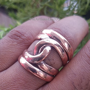 Op de afbeelding: Een brede koperen ring met een gepolijste, metalen afwerking en een in elkaar grijpend, geweven ontwerp dat om de vinger wikkelt. Het oppervlak van het metaal vertoont subtiele variaties in textuur en tint, met de tekst TheArtisanLeafUS zichtbaar.