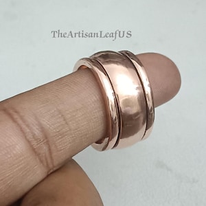 Peut inclure: Une large bague rotative en cuivre poli présentant une surface lisse et réfléchissante avec deux bandes extérieures plus fines. Le métal a une teinte brun-rouge chaude. Le texte TheArtisanLeafUS est visible dans la partie supérieure de l'image.