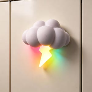 Könnte beinhalten: Eine weiße, wolkenförmige Wandleuchte mit strukturierter Oberfläche. Ein Blitzsymbol ragt nach unten heraus und strahlt ein lebendiges, mehrfarbiges Verlaufslicht aus, das in Gelb-, Orange-, Rosa- und Blautönen an der Wand schimmert.
