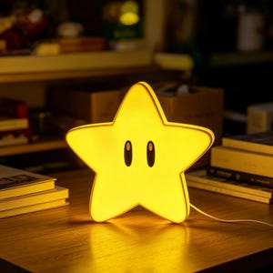 以下が含まれることがあります： 滑らかで光る表面を持つ、明るい黄色の星型ランプ。ランプの正面中央には、2つの大きな黒い楕円形の目が付いています。星型の照明器具の右下から、細い白い電源コードが伸びています。