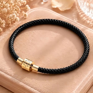 以下が含まれることがあります： 黒い編み込みブレスレットで、金色の金属製円筒形クラスプが付いています。クラスプにはP、O、N、D、O、O、Pの文字が円状に刻印されています。ブレスレットは均一な質感で、円形を保っています。