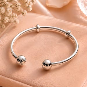 May include: A polished silver cuff bracelet featuring a smooth, open-ended circular band. The design includes two large, spherical end caps and two smaller, stationary ring-shaped beads positioned along the main body of the metallic bracelet.