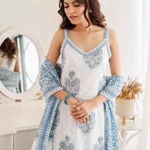 May include: A white sleeveless dress featuring a blue floral block print pattern, a V-neckline with blue trim, and a matching blue and white patterned shawl draped over the arms. The fabric has a subtle white dot texture throughout the garment.