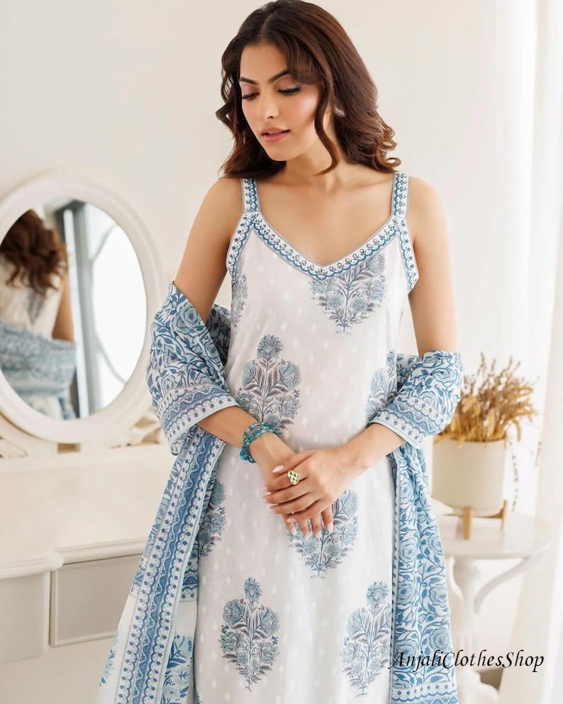 May include: A white sleeveless dress featuring a blue floral block print pattern, a V-neckline with blue trim, and a matching blue and white patterned shawl draped over the arms. The fabric has a subtle white dot texture throughout the garment.
