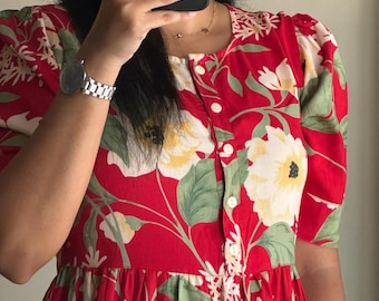 May include: A red dress featuring a floral pattern with large white and yellow flowers and green leaves. The dress has short puff sleeves, a round neckline, and a vertical row of white buttons extending from the collar down the center front.
