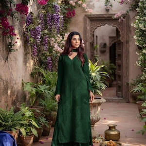 Puede incluir: Un vestido túnica largo de color verde oscuro con mangas largas, superficie de tela texturizada, escote en V con borde festoneado y cuello tipo gargantilla a juego, combinado con pantalones anchos del mismo tono y tejido.