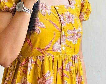 May include: A mustard yellow dress featuring a pink floral pattern, short puff sleeves, and a square neckline. The bodice includes a vertical row of four white buttons. The text AnjaliClothesShop is visible in the bottom right corner of the image.
