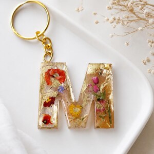 Può includere: Un portachiavi a forma di lettera M realizzato in resina trasparente con fiori secchi incorporati nei toni del rosso, giallo e rosa, arricchito da scaglie di foglia d'oro. La lettera è fissata a un anello in metallo dorato.