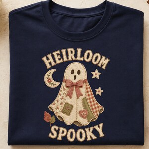 May include: A navy blue t-shirt featuring a graphic of a ghost made of patchwork fabric with a bow, surrounded by a crescent moon, stars, and autumn leaves. The text HEIRLOOM is arched above the ghost, and the word SPOOKY is printed below it.