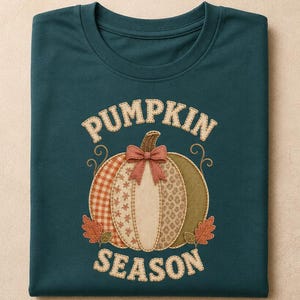 Puede incluir: Una camiseta de color verde azulado oscuro con un gráfico de una calabaza con varios patrones y un lazo. La calabaza está dividida en secciones con texturas de cuadros, flores y puntos. El texto PUMPKIN SEASON aparece en color crema arriba y abajo.