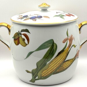 May include: A white porcelain lidded jar featuring gold-trimmed handles and a gold-accented lid knob. The body is decorated with a colourful illustration of corn on the cob and an apple, while the lid displays a pattern of small fruits and leaves.
