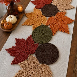 Può includere: Una collezione di decorazioni da tavola all'uncinetto con varie forme di foglie d'acero e sottobicchieri circolari in colori autunnali tra cui ruggine, marrone scuro, verde oliva e beige, disposti in fila su un runner da tavola beige chiaro.