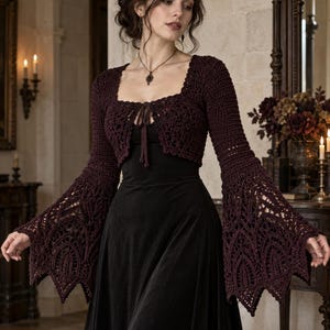 May include: A dark burgundy crochet bolero featuring long, flared bell sleeves with intricate lace-like patterns and a square neckline. The garment includes a front tie closure and is worn over a simple black dress, showcasing detailed needlework.