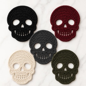 Puede incluir: Un conjunto de cinco posavasos de ganchillo con forma de calavera en negro, rojo oscuro, gris oscuro, crema y verde oliva. Cada posavasos presenta una forma de calavera detallada con cuencas oculares, una nariz triangular y una fila de dientes.
