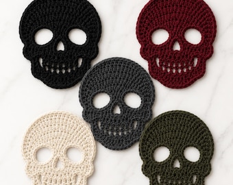 Puede incluir: Un conjunto de cinco posavasos de ganchillo con forma de calavera en negro, rojo oscuro, gris oscuro, crema y verde oliva. Cada posavasos presenta una forma de calavera detallada con cuencas oculares, una nariz triangular y una fila de dientes.
