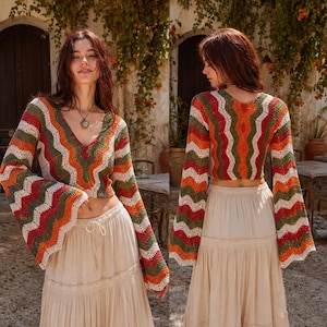 Pode incluir: Um top de crochê de manga comprida com um padrão chevron multicolorido em tons de laranja, verde e creme, com decote em V e mangas largas, combinado com uma saia longa de cor creme com camadas, cordão na cintura e detalhes em renda.