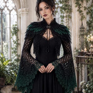 Puede incluir: Un bolero de ganchillo negro y verde oscuro con cuello alto, mangas largas acampanadas y un diseño de hombros en capas similar a plumas. La prenda se abrocha al cuello con un cierre decorativo y cordones finos sobre un vestido negro.