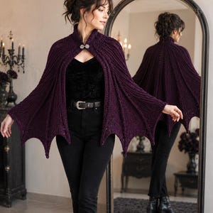 May include: A deep purple knitted cape featuring a ribbed texture and a scalloped, wing-like hemline. The garment is secured at the neck with a decorative silver-toned metal clasp. The knit pattern extends vertically from the collar to the hem.