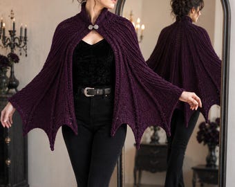 May include: A deep purple knitted cape featuring a ribbed texture and a scalloped, wing-like hemline. The garment is secured at the neckline with a decorative silver-toned metal clasp. The cape is worn over a black top and black trousers.