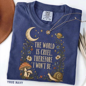 May include: A navy blue short-sleeve t-shirt featuring a graphic print of a crescent moon, a planet, mushrooms, flowers, and a snail. The text reads, "THE WORLD IS CRUEL, THEREFORE I WON'T BE." A thin gold-toned necklace rests on the collar area.