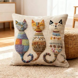 May include: A square beige throw pillow featuring three embroidered cats. The left cat has a multicolored patchwork pattern, the center cat is yellow with brown geometric details, and the right cat is white with floral and blue patterned accents.