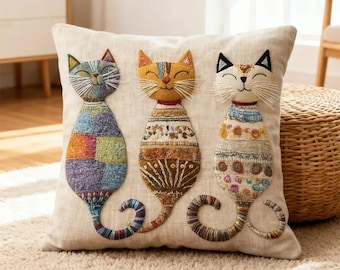 May include: A square beige throw pillow featuring three embroidered cats. The left cat has a multicolored patchwork pattern, the center cat is yellow with brown geometric details, and the right cat is white with floral and blue patterned accents.