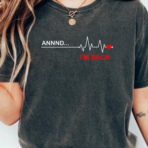 May include: A charcoal gray short-sleeve t-shirt featuring the white text ANNND... followed by a white EKG heartbeat line that ends in a small red heart icon, with the red text I'M BACK positioned directly underneath the heartbeat graphic.