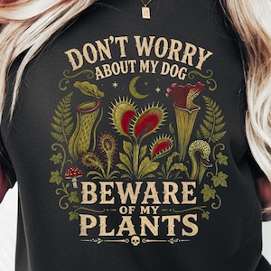 May include: A black t-shirt featuring a graphic print of various carnivorous plants, including a Venus flytrap and pitcher plants. The text reads, "DON'T WORRY ABOUT MY DOG BEWARE OF MY PLANTS," accompanied by small illustrations of a crescent moon, a mushroom, and a skull.